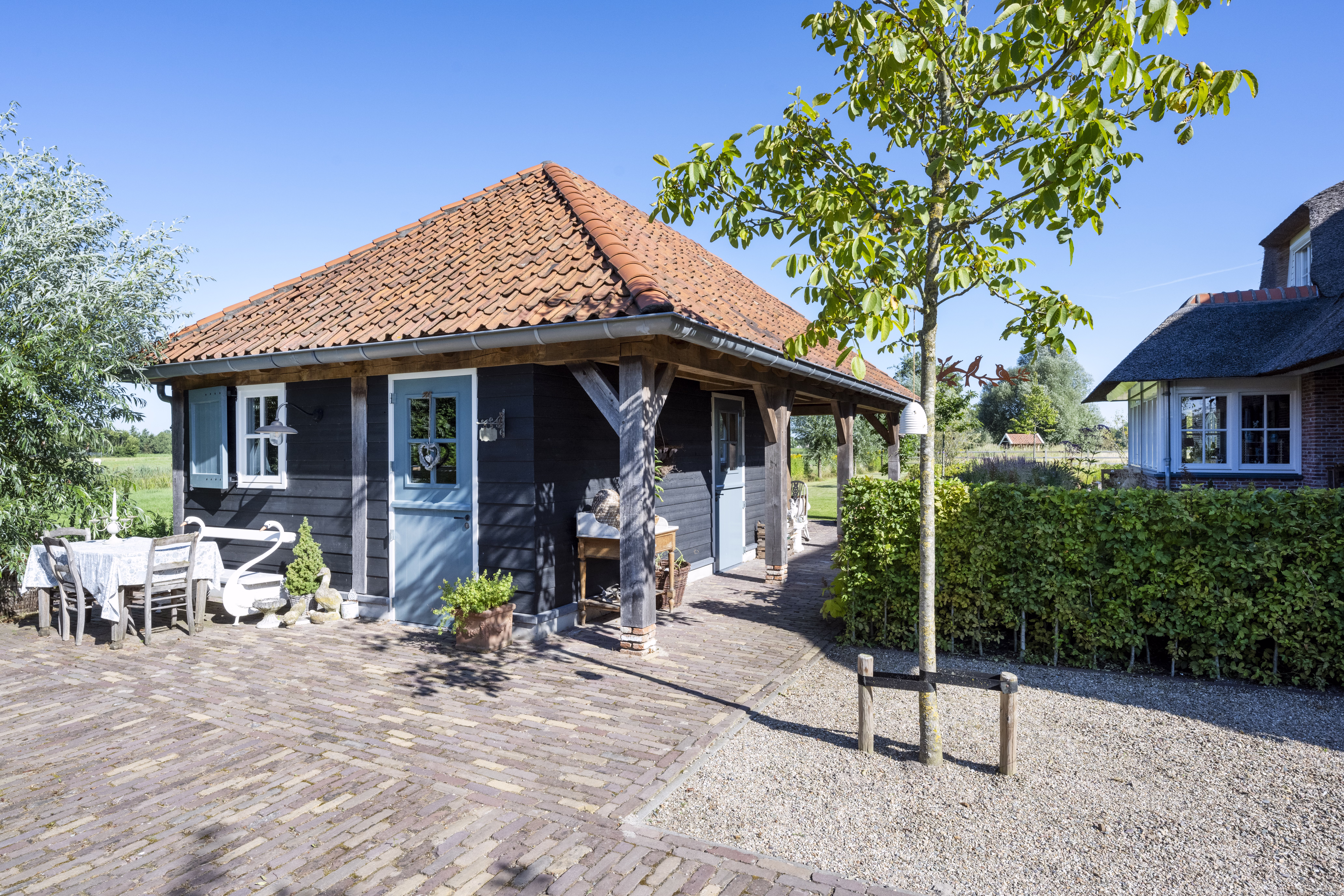 Bijgebouw met zwart houten gevelbekleding, rode dakpannen en witten kozijnen en blauwe luiken passend bij de woning.